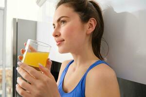 dichtbij omhoog portret van mooi jong vrouw, op zoek weg, drinken oranje sap, vers in glas, staand in keuken, vervelend training kleren foto