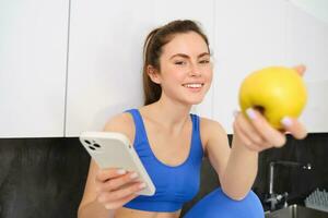 beeld van mooi, sportvrouw zittend in keuken met smartphone, aanbieden u een appel, aan het eten fruit, vervelend sport bh en leggings foto