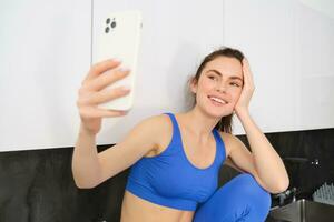 beeld van fit en gezond, glimlachen geschiktheid meisje, poseren voor selfie Aan mobiel telefoon, Holding smartphone, op zoek Bij scherm, nemen foto's in sport bh en leggings foto