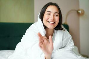 gelukkig mooi Aziatisch vrouw gedekt met deken, zit Aan bed in warm donsdeken, probeert naar bereiken camera met een hand, glimlachen speels. hotel slaapkamer, comfort en appartementen concept foto