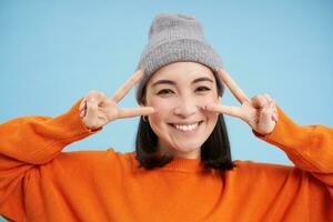 dichtbij omhoog portret van schattig Koreaans meisje in muts, shows vrede, vsign gebaar, positief uitstraling, glimlachen en lachend, poseren over- blauw achtergrond foto