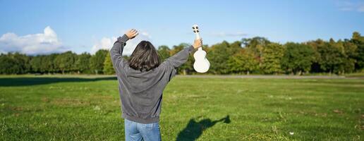 vrolijk jong vrouw dansen met haar musical instrument. meisje verhoogt haar ukulele omhoog en houding in park Aan groen veld- foto