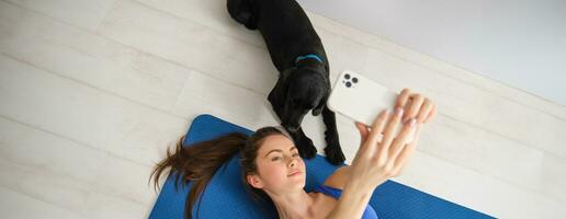 portret van mooi sportvrouw, geschiktheid meisje aan het liegen Aan verdieping Bij huis met haar hond, nemen selfie met pup, vervelend training kleren foto