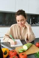 verticaal schot van glimlachen vrouw maken notities, schrijven Koken recept, creëert boodschappen lijst, zit in de keuken met groenten en hakken bord foto