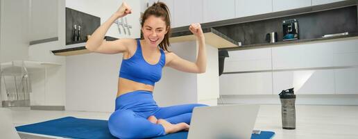 beeld van opgewonden jong vrouw tonen spieren, buigen biceps en op zoek Bij laptop scherm, training van huis, geschiktheid instructeur onderwijs van een afstand, geven online yoga les foto