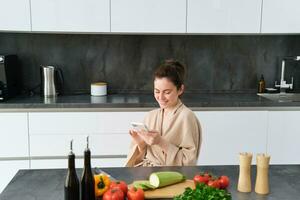 vrouw aan het kijken recept Aan smartphone, zittend in de keuken met groenten en hakken bord, voorbereidingen treffen diner, gezond salade, Koken Bij huis, vervelend badjas foto