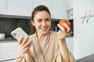 beeld van glimlachen mooi vrouw, zit in keuken, houdt smartphone en tomaat, looks Vrolijk, bestellingen boodschappen levering naar haar huis, gebruik makend van mobiel telefoon app, buying online groenten voor haar recept foto