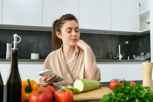 portret van jong vrouw bestellen boodschappen Aan smartphone app, Holding tomaat, zittend in de buurt hakken bord met groenten. meisje op zoek voor recept online, gebruik makend van mobiel telefoon foto