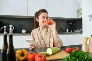 portret van jong vrouw bestellen boodschappen Aan smartphone app, Holding tomaat, zittend in de buurt hakken bord met groenten. meisje op zoek voor recept online, gebruik makend van mobiel telefoon foto