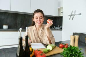 portret van mooi glimlachen vrouw, schrijven haar gezond menu, aan het eten tomaat terwijl Koken, maken kruidenier lijst, zittend in de keuken foto