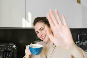 portret van schattig jong vrouw, covers haarzelf van camera, lachend en flirten, drinken koffie in de ochtend, staand in de keuken foto