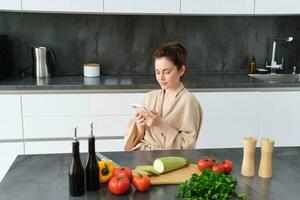 vrouw aan het kijken recept Aan smartphone, zittend in de keuken met groenten en hakken bord, voorbereidingen treffen diner, gezond salade, Koken Bij huis, vervelend badjas foto