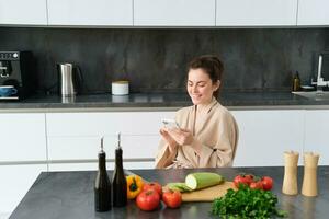 portret van vrouw in badjas zittend in keuken met smartphone, Koken diner, aan het kijken recept Aan sociaal media, video zelfstudie hoe naar bereiden maaltijd foto