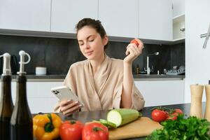 beeld van jong mooi vrouw, Holding tomaat, zittend in keuken met smartphone, hakken bord en groenten Aan balie, Koken voedsel, bestellen boodschappen voor haar recept, gebruik makend van mobiel telefoon app foto