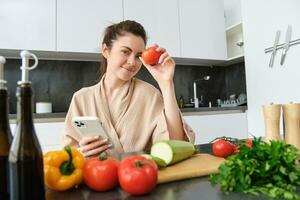 beeld van jong mooi vrouw, Holding tomaat, zittend in keuken met smartphone, hakken bord en groenten Aan balie, Koken voedsel, bestellen boodschappen voor haar recept, gebruik makend van mobiel telefoon app foto