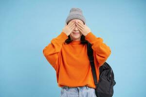 schattig Aziatisch vrouw met rugzak, sluit ogen en wacht voor verrassing, staat over- blauw achtergrond foto