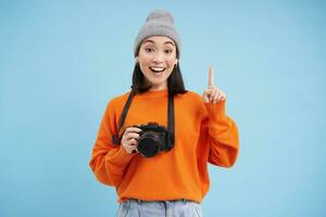 verrast Aziatisch meisje in hoed, houdt digitaal camera, looks versteld staan en onder de indruk, poses over- blauw studio achtergrond met ruimte voor banier foto