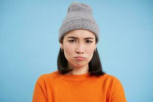 dichtbij omhoog portret van mokkend, verdrietig Aziatisch vrouw in winter hoed, fronsen en op zoek boos, beledigd gezicht, staand over- blauw achtergrond foto