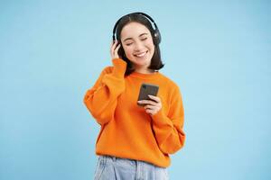 gelukkig jong modern vrouw met hoofdtelefoons en smartphone, luisteren naar muziek- Aan mobiel telefoon, geniet favoriete podcast, blauw achtergrond foto