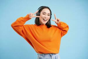 mooi Koreaans vrouw in koptelefoon, dansen en luisteren muziek- in oortelefoons, staand over- blauw achtergrond foto