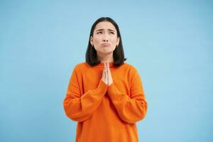 bidden naar god. ellendig Aziatisch meisje houdt handen in smeekbede, maakt wens, looks omhoog en wacht voor wonder, staat over- blauw achtergrond foto