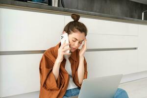 portret van bezorgd vrouw, zit Aan verdieping met laptop, luistert naar slecht nieuws over- de telefoon, roeping iemand met van streek, bezorgd gezicht uitdrukking foto