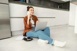 portret van glimlachen vrouw met smartphone, pratend Aan mobiel telefoon, roeping iemand terwijl zittend Aan verdieping Bij huis met laptop foto