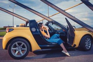 jong meisje in blauw jeans, oranje top en omdraaien flops is aan het eten Hamburger terwijl zittend in geel auto cabrio. snel voedsel, ongezond voeding foto