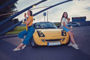 Dames in gewoontjes kleding zijn genieten van hamburgers, Holding dranken in papier cups terwijl zittend Aan de kap van geel auto. snel voedsel. kopiëren ruimte foto