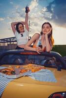 gelukkig Dames hebben plezier, drinken Frisdrank water in glas flessen, poseren in geel auto cabriolet met Frans Patat en pizza Aan kofferbak. dichtbij omhoog foto