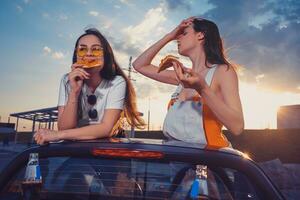 gelukkig modellen in gewoontjes kleding zijn aan het eten pizza, poseren in geel auto roadster met Frisdrank water in glas flessen Aan haar kofferbak. snel voedsel. kopiëren ruimte foto