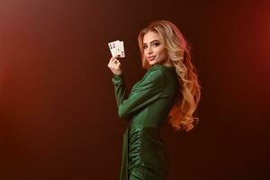 blond dame in groen jurk en sieraden. lachend, op zoek Bij jij, tonen twee azen, poseren zijwaarts Aan bruin achtergrond. poker, casino. detailopname foto