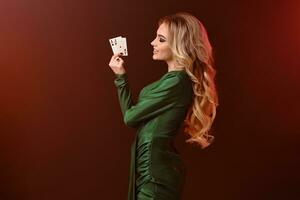 blond meisje in groen jurk en sieraden. lachend, gevouwen haar handen, tonen twee azen, poseren zijwaarts Aan bruin achtergrond. poker, casino. detailopname foto