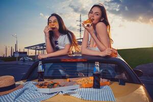 charmant vrouwtjes zijn aan het eten pizza terwijl poseren in geel auto cabriolet met Frans Patat en Frisdrank in glas flessen Aan kofferbak. snel voedsel. dichtbij omhoog foto