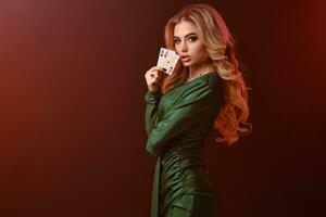 blond model- in groen elegant jurk en sieraden. gevouwen handen, tonen twee spelen kaarten, poseren zijwaarts Aan rood achtergrond. poker, casino. detailopname foto
