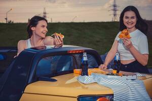 mooi Dames lachend, aan het eten pizza, poseren in geel auto cabrio met Frans Patat en Frisdrank in glas flessen Aan haar kofferbak. snel voedsel. kopiëren ruimte foto