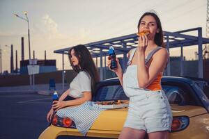 aantrekkelijk Dames in gewoontjes kleren aan het eten pizza, genieten van Frisdrank water in glas flessen, poseren in de buurt geel auto Aan parkeren kavel. dichtbij omhoog, kopiëren ruimte foto