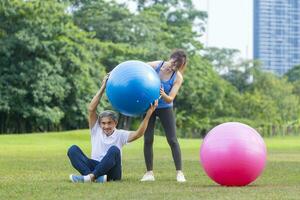 senior Aziatisch Mens en zijn ondersteunen dochter zijn oefenen met yoga bal in openbaar park naar bouwen kern lichaam spier voor ouderling levensduur oefening en buitenshuis training foto