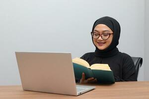 portret van aantrekkelijk Aziatisch hijab vrouw werken Aan haar laptop. moslim meisje lezing een boek in kantoor. werknemer en freelance arbeider concept. foto