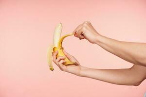 studio schot van elegant vrouw handen Holding banaan terwijl pellen het en gaan naar beet, hebben gezond ontbijt terwijl wezen geïsoleerd over- roze achtergrond foto