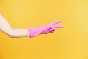 studio foto van jong vrouw hand- in rubber handschoen vormen zege gebaar met twee vingers terwijl wezen geïsoleerd over- oranje achtergrond. tekens en gebaren concept