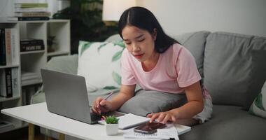 portret van jong Aziatisch vrouw cheques haar financiën gebruik makend van een rekenmachine Aan smartphone en maakt aantekeningen in een notitieboekje terwijl zittend Aan de sofa Bij huis huis financiën foto