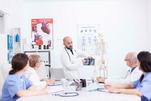 jong dokter geven presentatie naar collega's in conferentie kamer Holding röntgenfoto. kliniek deskundige therapeut pratend met collega's over ziekte, geneeskunde professioneel. foto