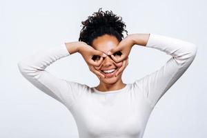 portret van een positieve en emotionele mooie jonge vrouw, gemaakt van vingers een figuur vergelijkbaar met de pilootbril rond haar ogen geïsoleerd op een witte muur. foto