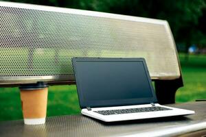 mockup beeld van laptop met blanco zwart scherm en koffie kop Aan metaal bank in natuur buitenshuis park foto