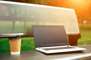 mockup beeld van laptop met blanco zwart scherm en koffie kop Aan metaal bank in natuur buitenshuis park foto