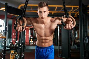 portret van jong Mens gebruik makend van gymnastiek- ringen terwijl oefenen in Sportschool foto