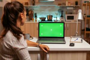 vrouw op zoek Bij laptop met groen mockup gedurende nacht tijd in huis keuken. zittend Bij bureau werken Aan computer laat Bij nacht, bedrijf, online, slim, blanco, kopieerruimte. foto