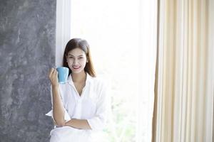 Aziatische vrouwen die koffie drinken en volledig uitgerust wakker worden in haar bed en 's ochtends de gordijnen openen om frisse lucht te krijgen in de zon foto