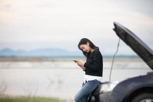 Aziatische vrouw die mobiele telefoon gebruikt terwijl ze kijkt en gestresste man zit na een autopech op straat foto